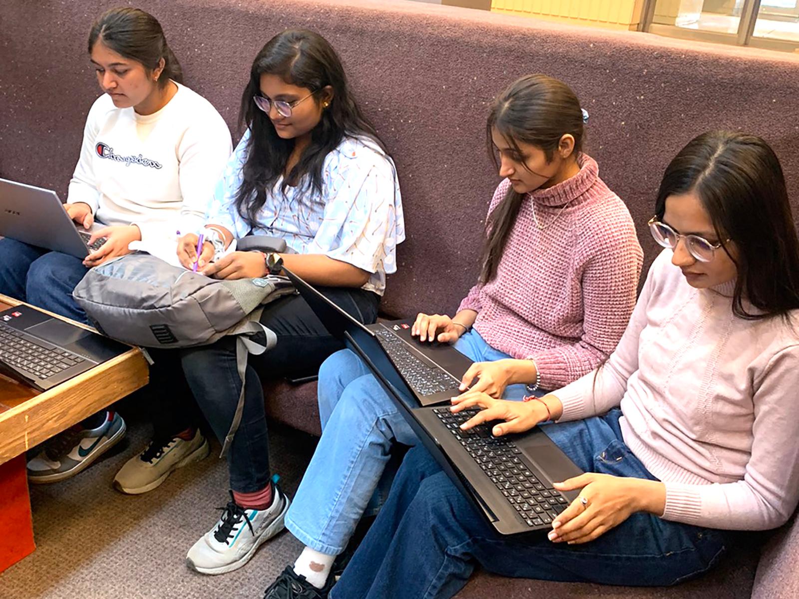 four female students sitting in a corner of a large brown bench, three are looking at open laptops, the fourth is writing on a piece of paper