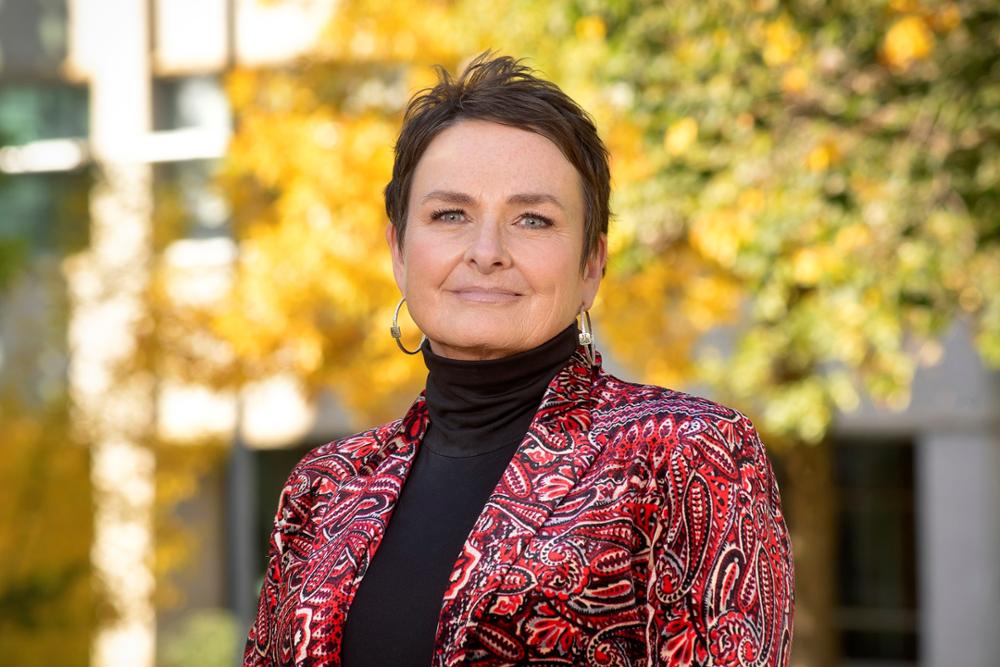 : In a close-up outdoor picture, Chancellor Pam Klein is looking directly at the camera with trees in fall colours and a building in the background 