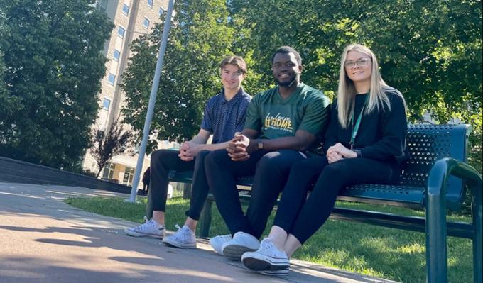 Three students sitting on a bench outdoors with trees and the Paskwā and Wakpāw residence towers in the background. 