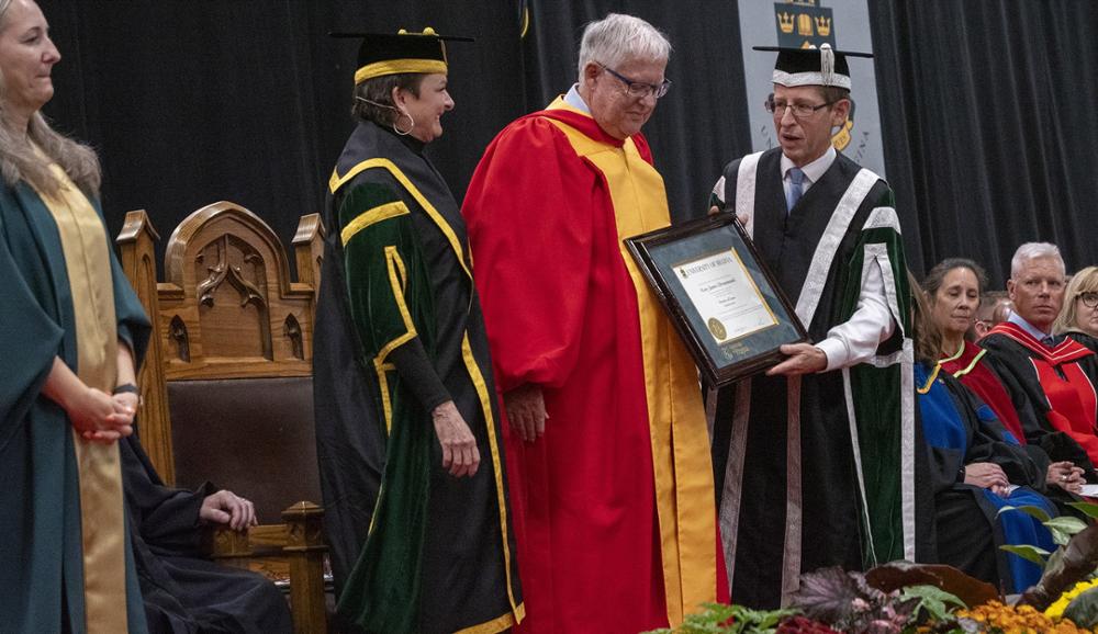Three people on stage, one is receiving an honourary degree.