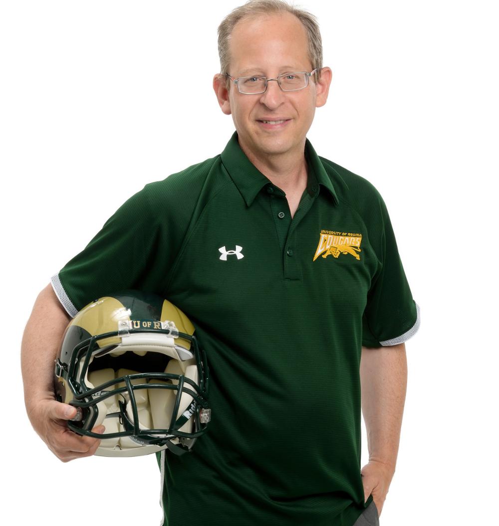 U of R President Jeff Keshen wearing Cougars gear holding a U of R Rams helmet.