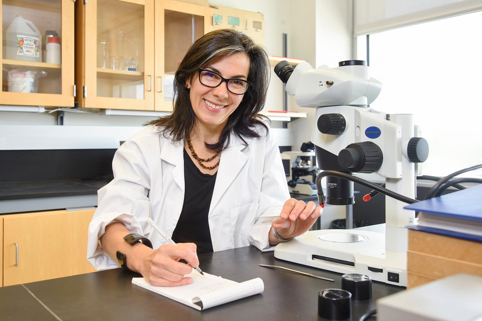 A professor at the head of a classroom leaning over a table and smiling at the camera.