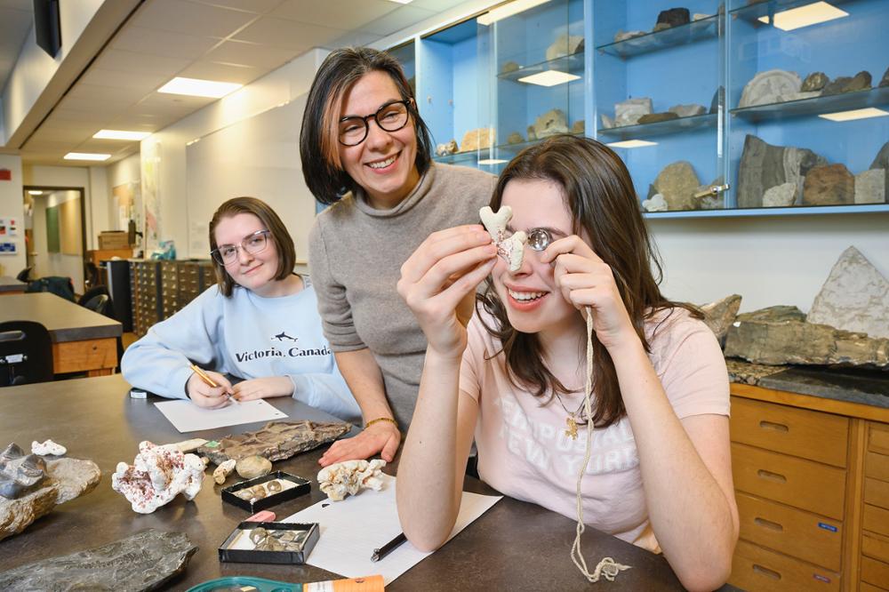 Professor and two students in a lab with one student examining fossil through magnifying glass.