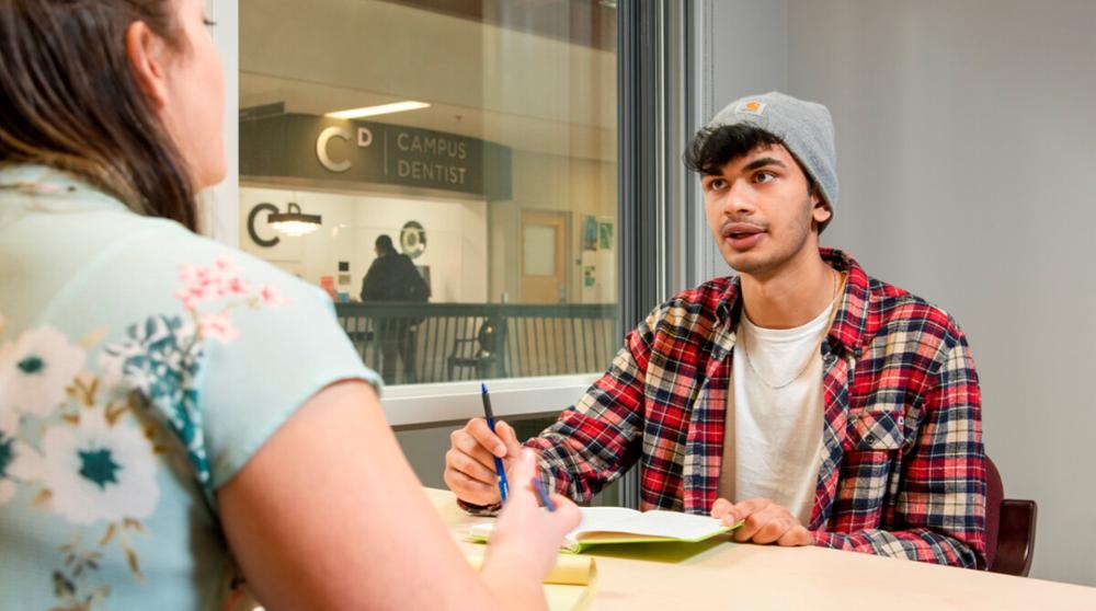 Male student and advisor seated across from one another at a desk and talking.