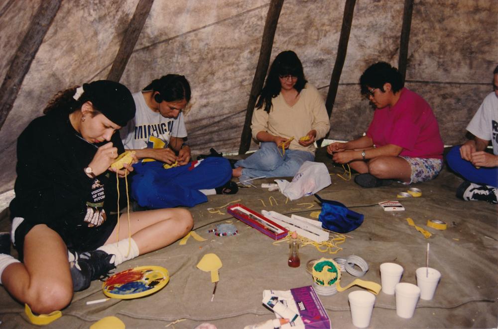 Five individuals sitting inside of a tipi, creating beadwork designs.