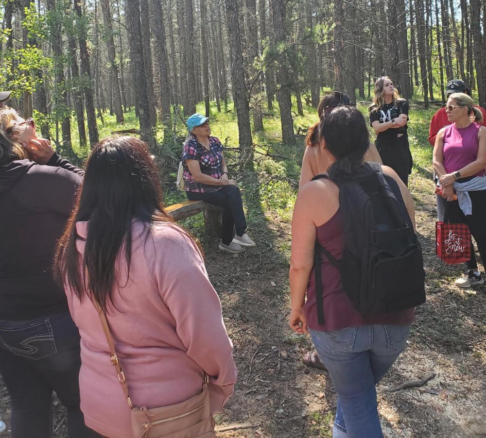 Woman sitting on bench in forest speaking to a group of people standing on path.