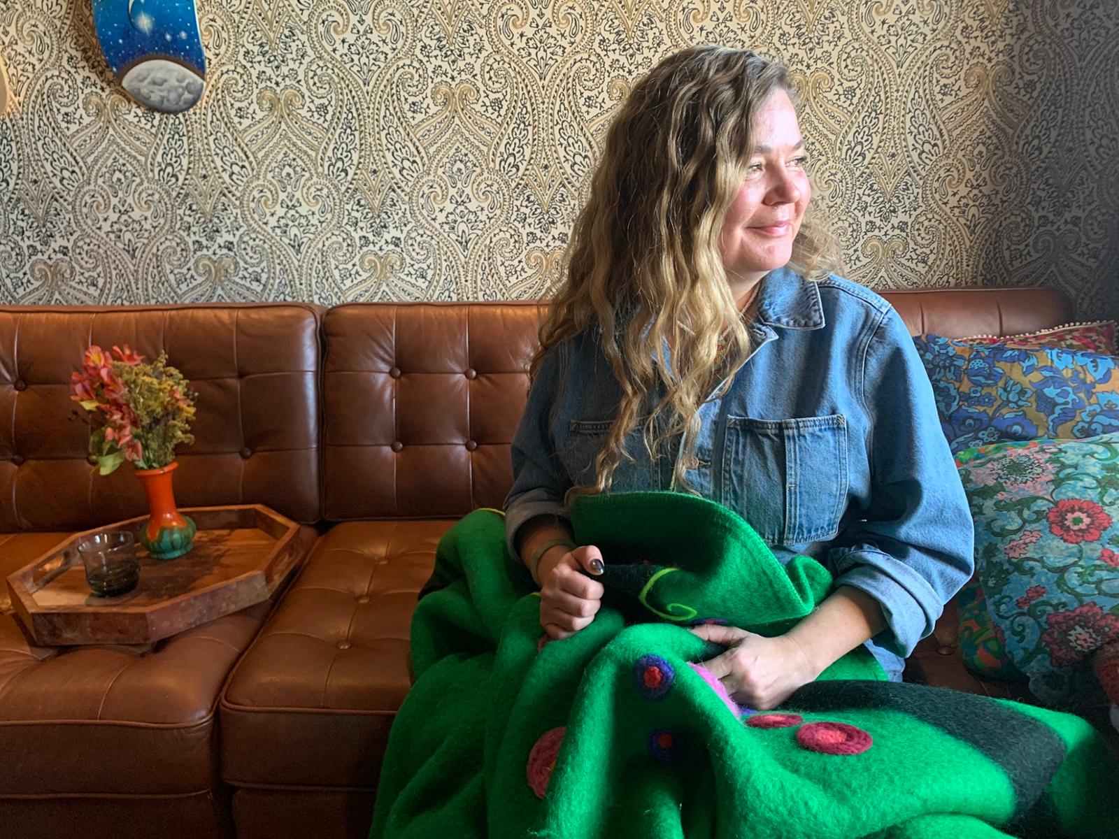 Woman sits on a couch with an ornate blanket on her lap. 