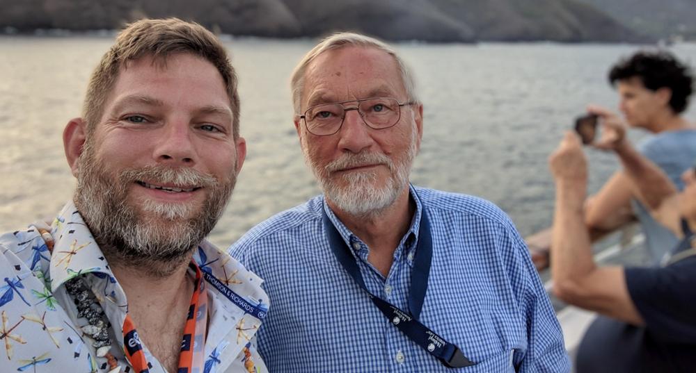 Photo of two men standing on the deck of a ship with mountains behind them