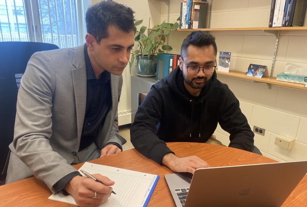 Two men sit at office desk looking down at a laptop that is open.