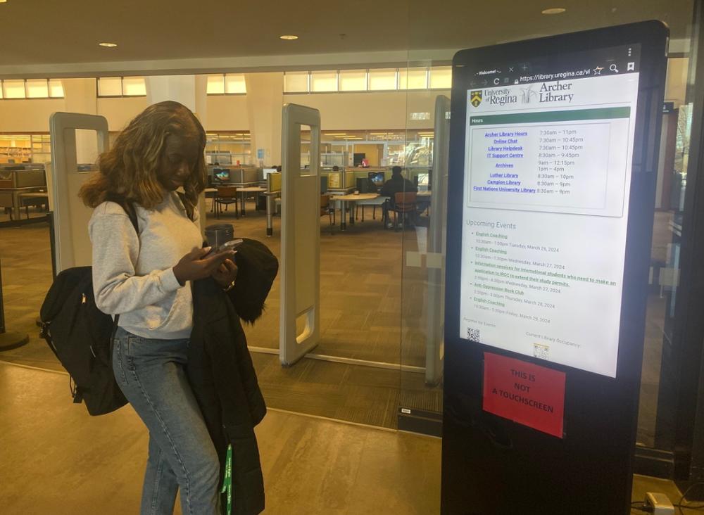 Female student standing looking at her mobile phone beside digital kiosk at the Library entrance. 