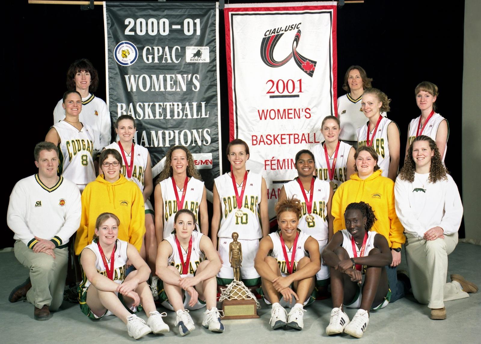 Women basketball team poses for photo in front of 2000-01 banners highlighting winning season.
