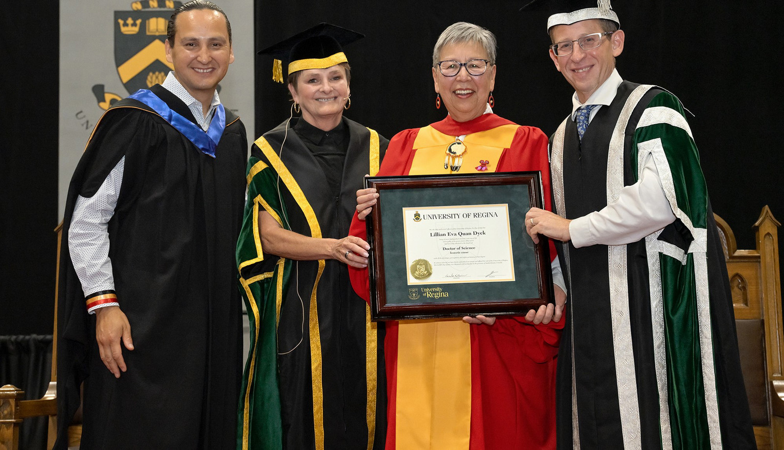 Four people in ceremonial robes standing close together and smiling while one holds a framed parchment.