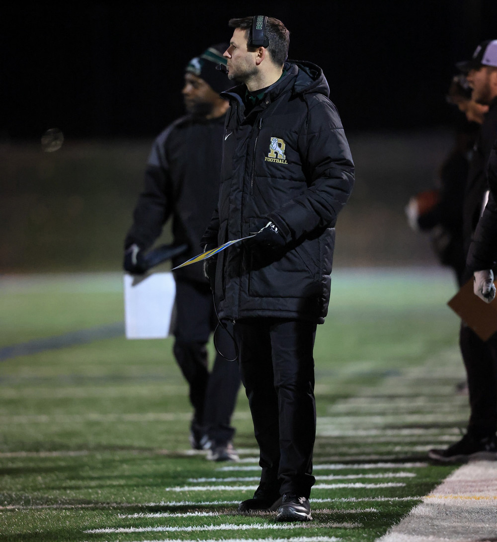 A man with headset on watches a football game from the sidelines.