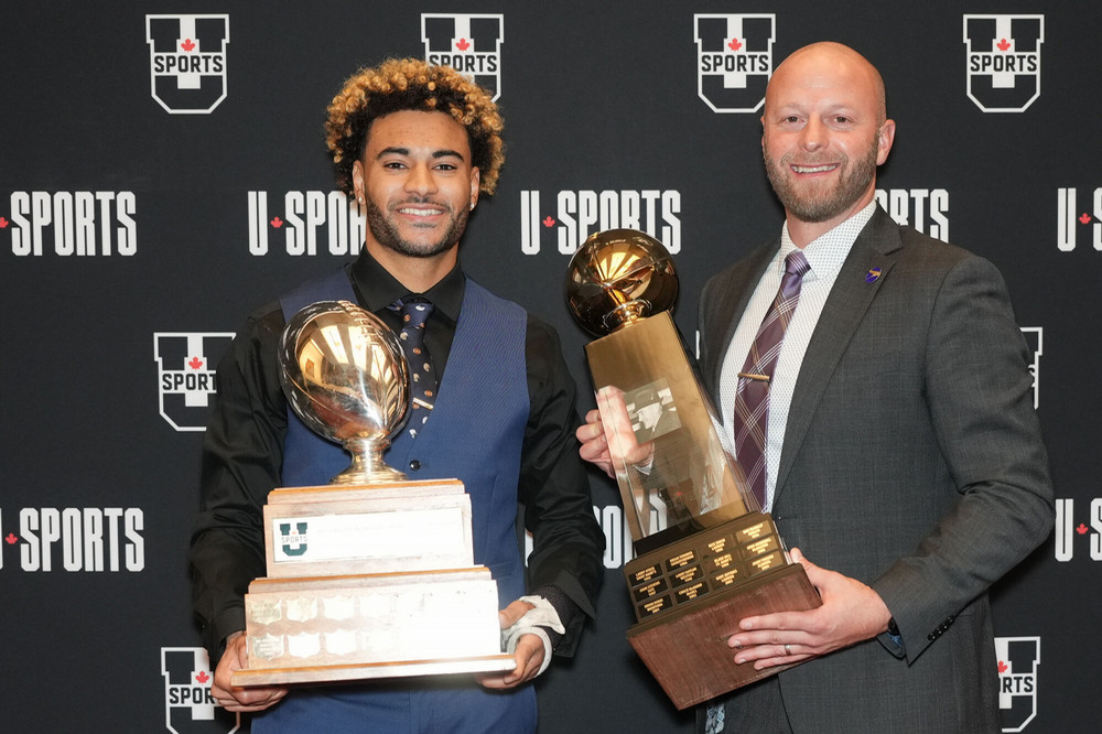 A young man and an older man pose with large trophies. 
