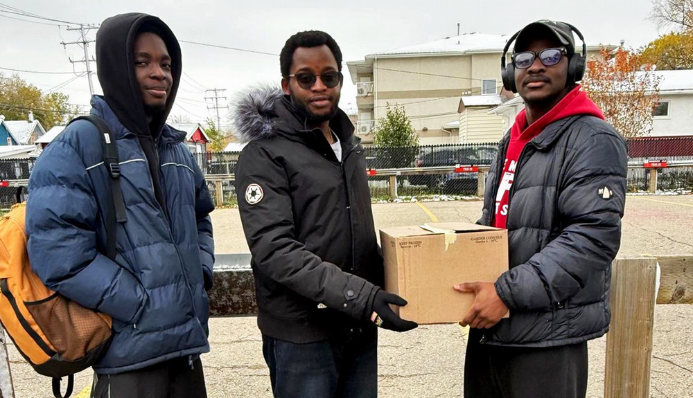 Two people face the camera holding a box and a third looks on wearing a backpack. 
