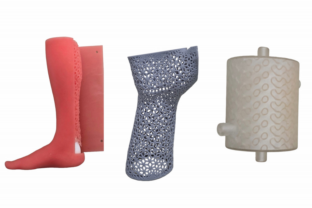 A side-by-side display of three 3D-printed research prototypes: a pink colored lower-leg prosthetics model showing internal structural density (left), a grey lattice-patterned orthotics model (centre), and a translucent white cylindrical heat exchanger prototype with complex internal channels (right).