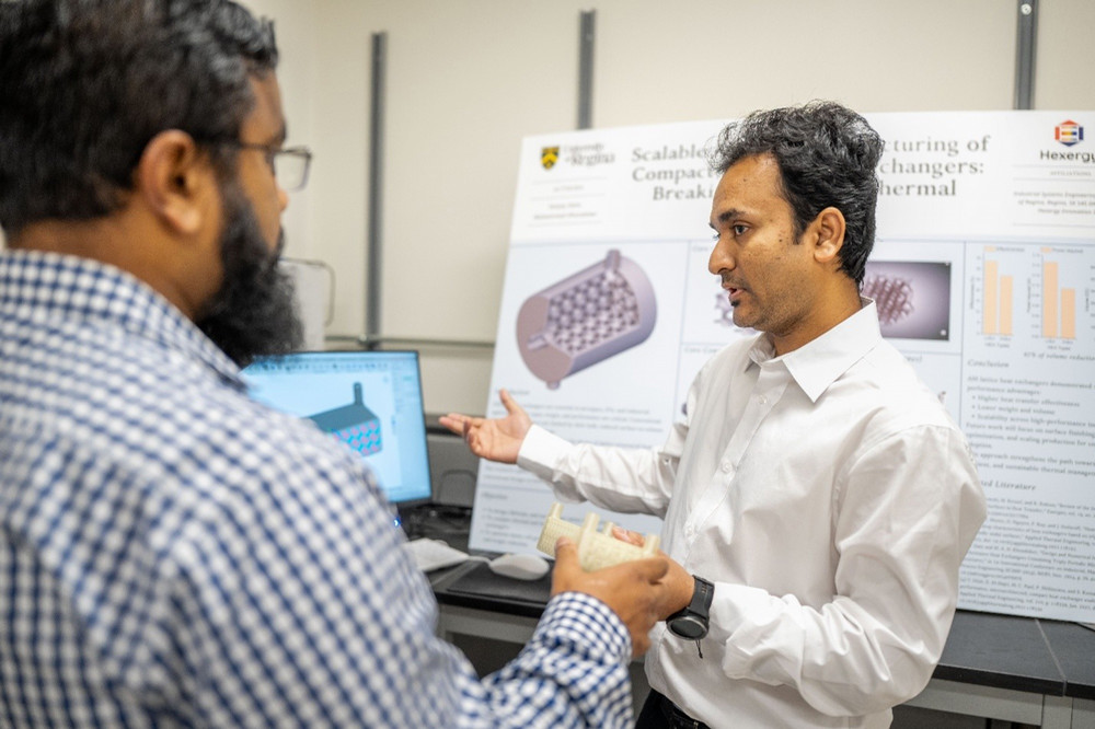Two researchers standing in a lab discuss a beige, 3D-printed component held by one of them, with a technical poster titled "Scalable Additive Manufacturing of Compact Heat Exchangers" visible in the background.