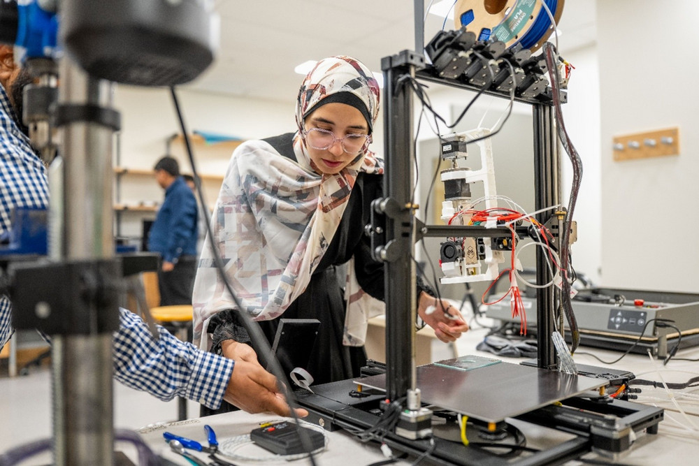 A close-up shot of a researcher in a lab coat and hijab meticulously adjusting the components of a complex 3D printer, surrounded by laboratory equipment.