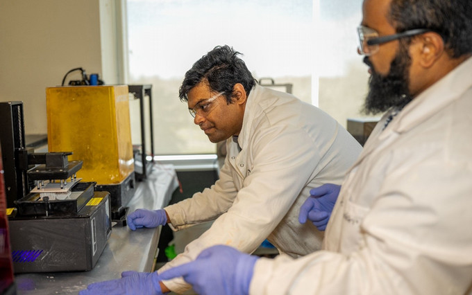 A side-view shot of two researchers in white lab coats examining a small, yellow 3D printer in operation, focusing intently on the printing process.
