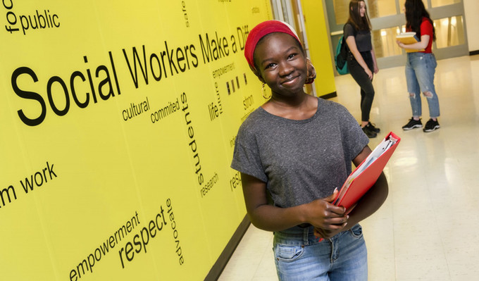 A young woman student stands in front of a Social Work sign. 