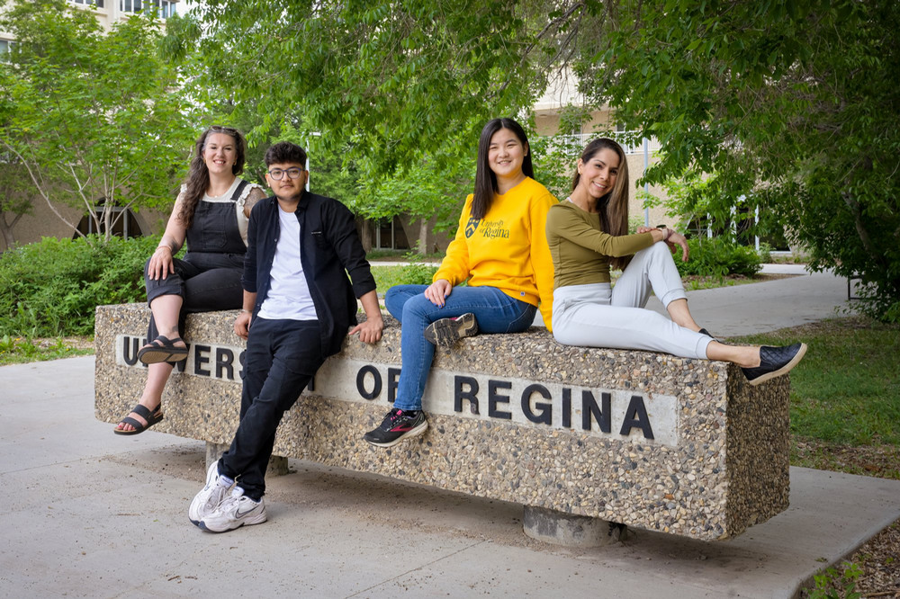 A diverse group of four smiling students sitting together casually on a concrete University of Regina sign in a leafy outdoor campus setting.