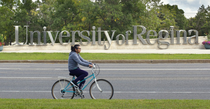 A cyclist rides past the large, silver University of Regina sign on a sunny day, with lush green trees and a blue sky in the background at the Wascana Park campus.