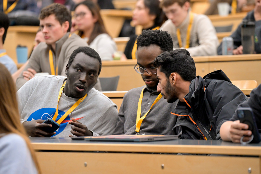 Three students wearing yellow lanyards sit together in a lecture hall, leaning in and discussing ideas