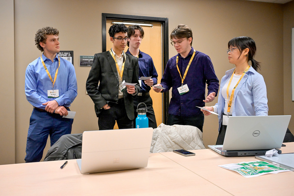 Five students stand in a breakout room discussing their ideas, with laptops and notes on a table 