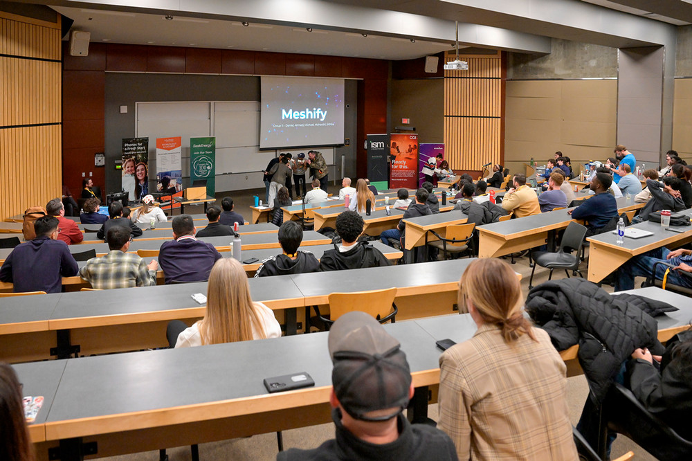 Wide view of a lecture hall, with students seated and a team presenting at the front 