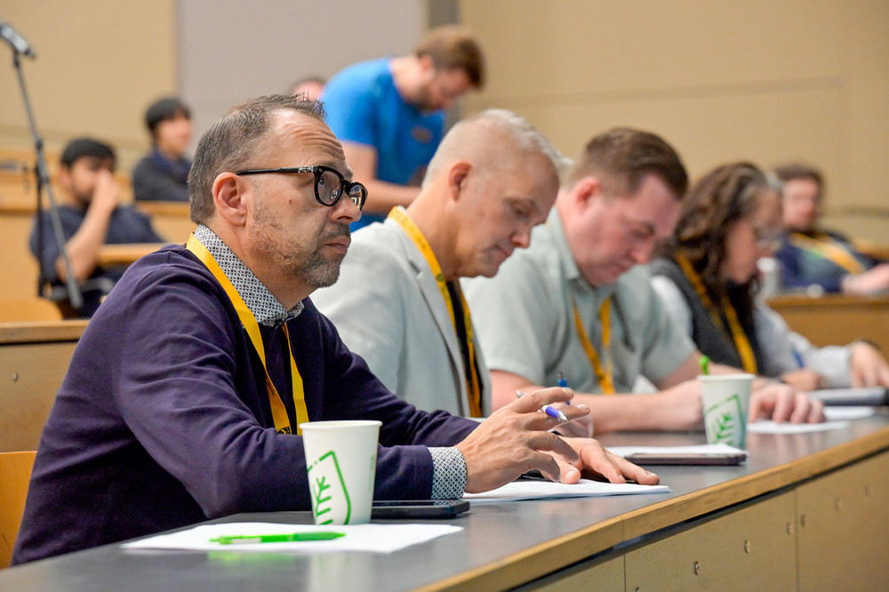Three Crown partner judges wearing yellow lanyards sit at a table in a lecture hall, listening and taking notes 