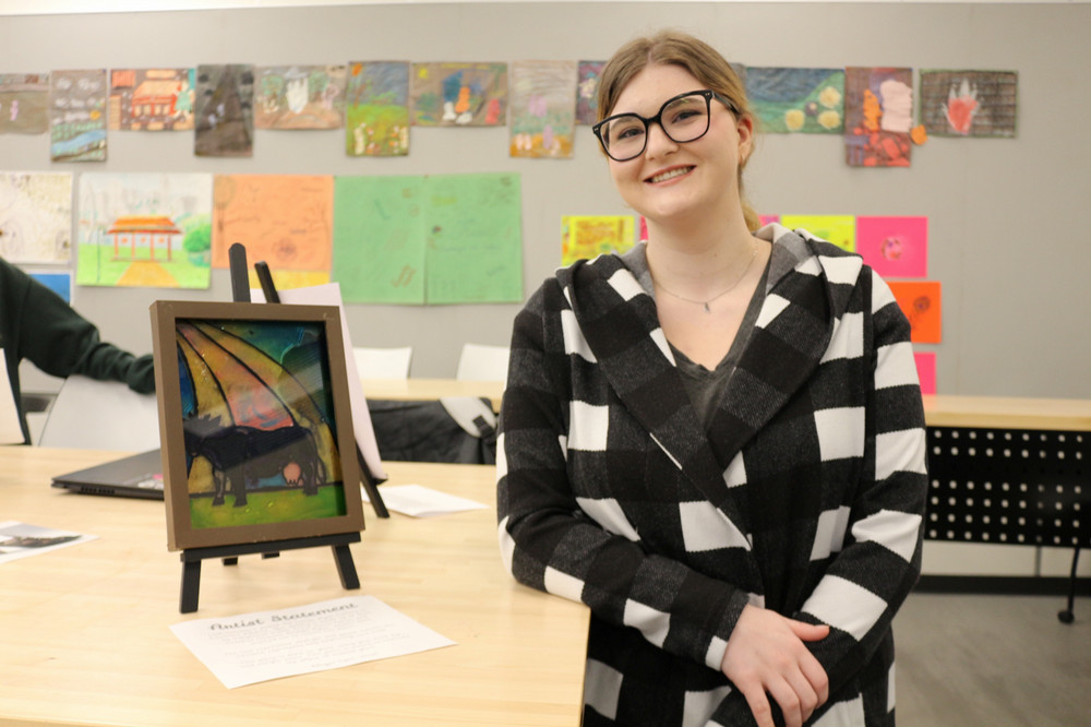 A student stands beside a small, framed artwork displayed on a tabletop easel in a classroom. Colourful student artwork is displayed on the wall behind them.