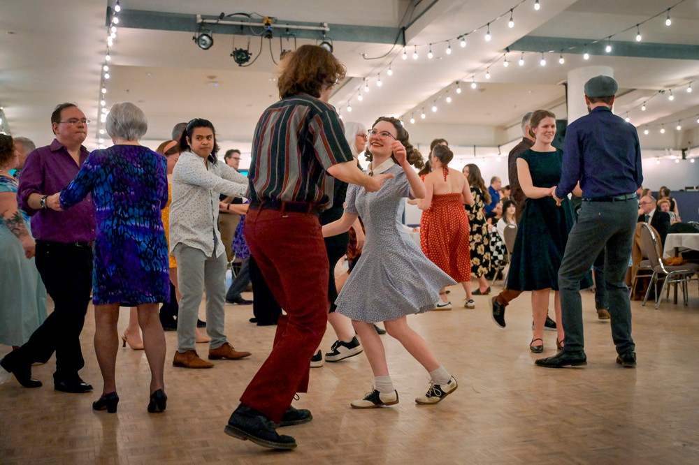 People swing dance together on a crowded dance floor under string lights during a big band jazz event.