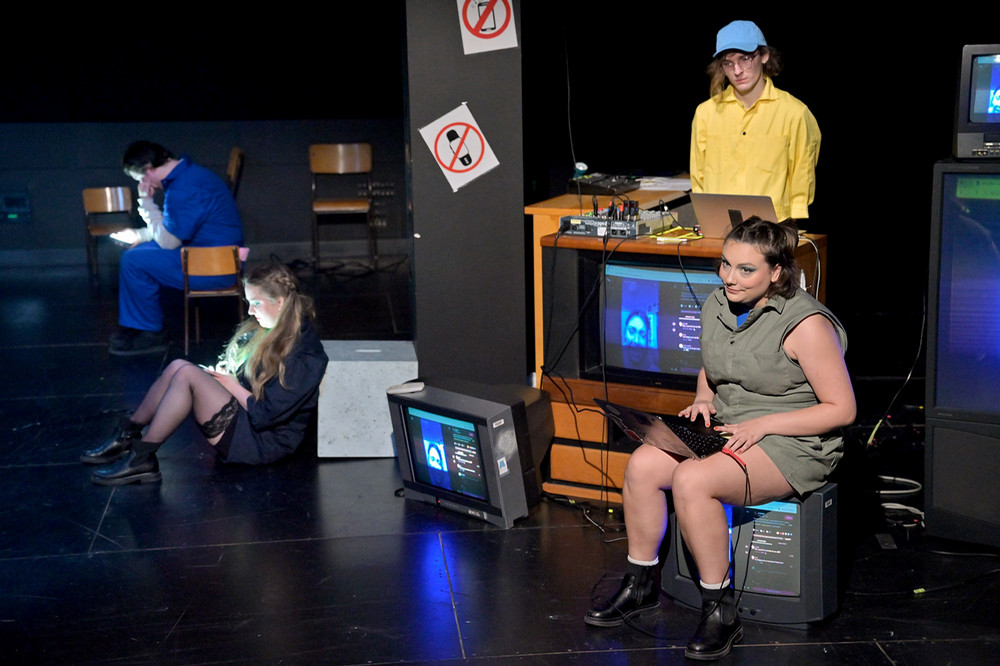 Several performers are spaced across a dark stage, seated or standing near televisions, laptops, and audio equipment, with glowing screens throughout.