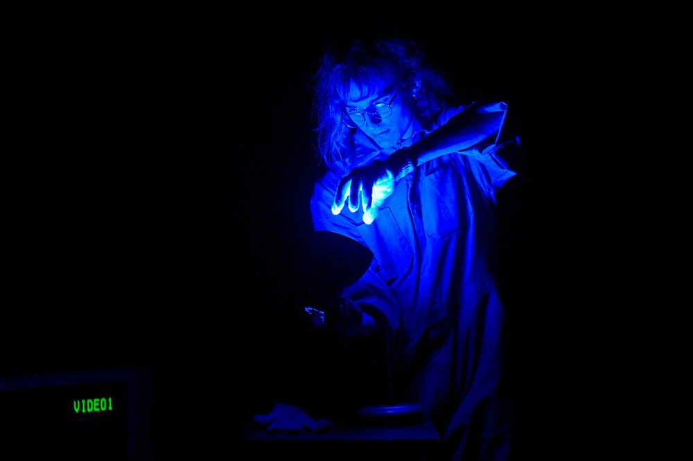  A performer is illuminated in blue light while leaning over a table and holding their hand over a light. 