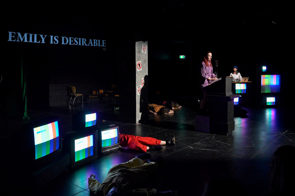 A wide stage scene shows two performers standing at podiums while several other performers lie motionless across the stage floor. Multiple televisions display color bars, and large projected text reading “EMILY IS DESIRABLE” appears on the back wall. 