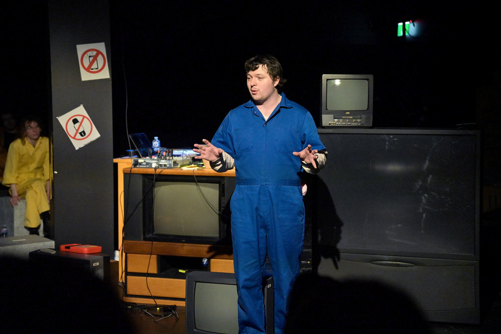 A performer stands on stage wearing blue coveralls, hands raised mid‑gesture. Behind the performer are stacked televisions, stage equipment, and a small table with electronics. Another performer in a yellow outfit is partially visible in the background.