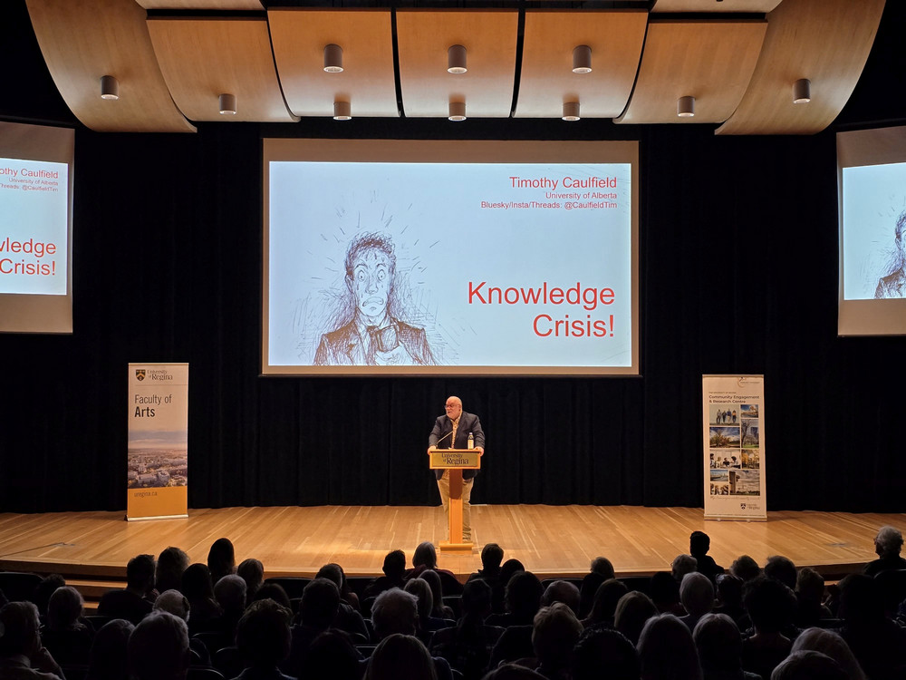 Associate Dean Tom McIntosh addresses a capacity audience from the University of Regina podium during the 2026 Woodrow Lloyd Lecture, with Timothy Caulfield's Knowledge Crisis title slide projected behind him.