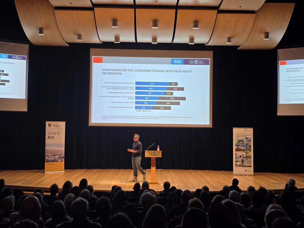 Timothy Caulfield presents survey data on Canadian health misinformation beliefs during the 2026 Woodrow Lloyd Lecture at the University of Regina, with a capacity audience visible in the foreground. 