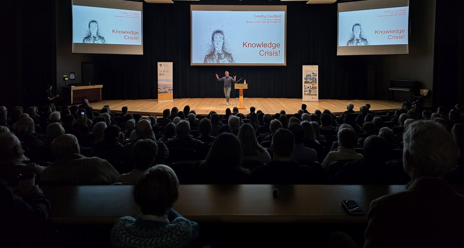 A wide-angle view of the packed Education Auditorium during the 2026 Woodrow Lloyd Lecture, with Timothy Caulfield on stage addressing a capacity crowd and 'Knowledge Crisis!' displayed across three screens
