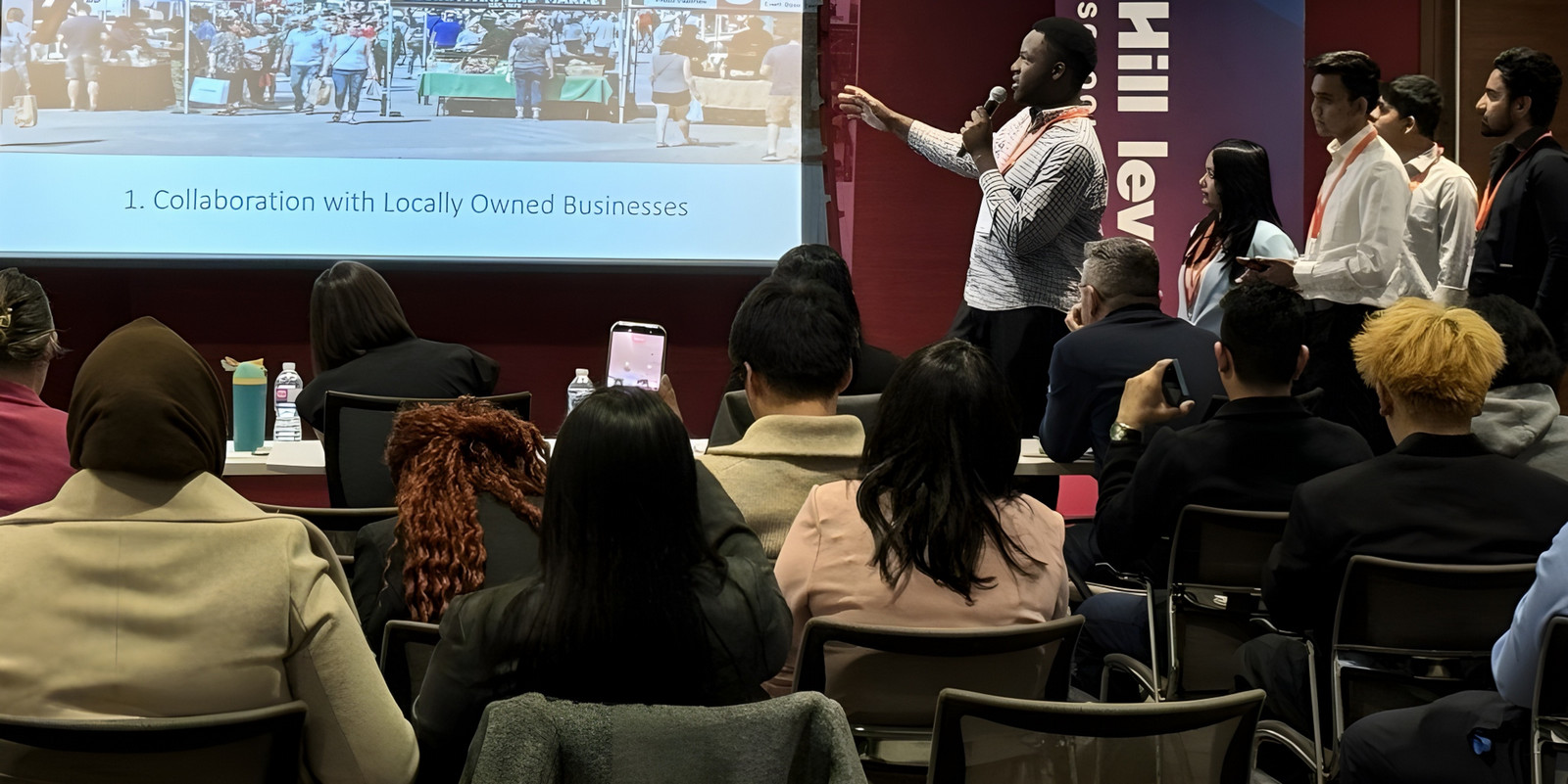 student gestures toward a projector screen displaying a mock-up of a revitalized downtown market, while teammates stand nearby, presenting to a full room of judges, peers, and industry mentors