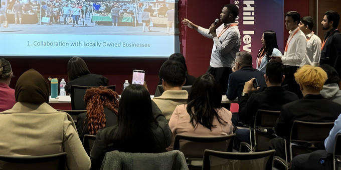 student gestures toward a projector screen displaying a mock-up of a revitalized downtown market, while teammates stand nearby, presenting to a full room of judges, peers, and industry mentors