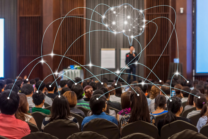 Audience members seated in a conference hall facing a speaker on stage
