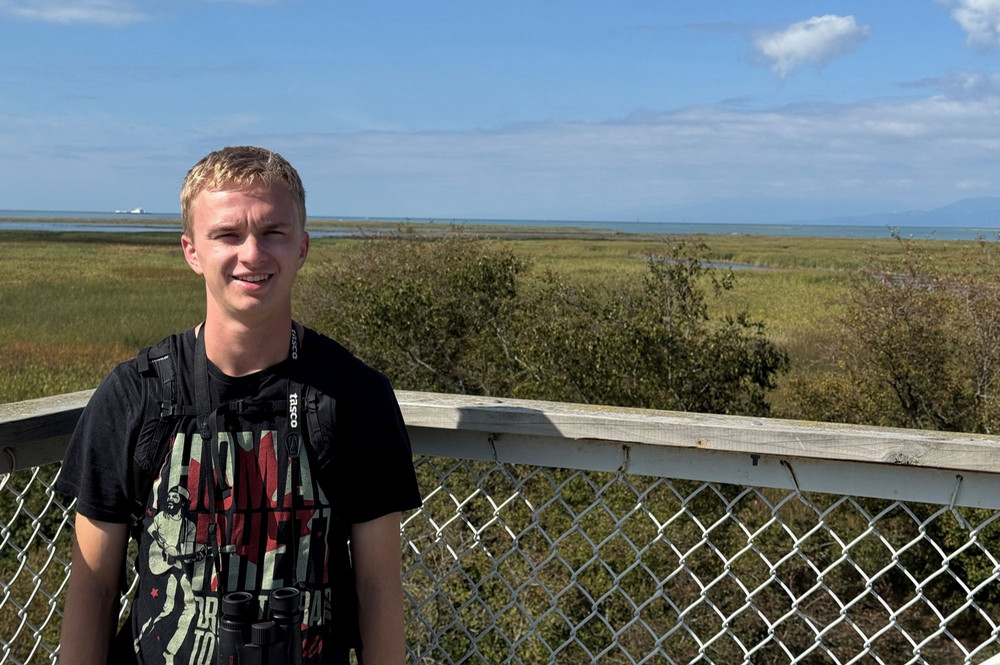 person standing in front of a chain-link railing, wearing a black graphic T-shirt and binoculars, with a wide grassy wetland and blue sky in the background.