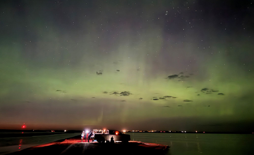 Nighttime view of the northern lights glowing green and purple across a star-filled sky above dark water, with a truck in the foreground illuminated by red and white lights.