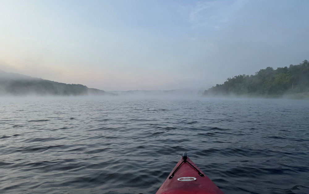View from the front of a red kayak on a lake, with gentle ripples on the water, low mist drifting across the surface, and tree-lined shores on both sides under a pale blue sky.