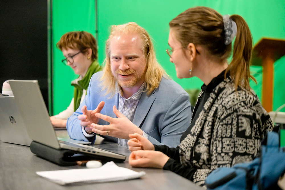 A man sits beside a young woman in front of a laptop in a university classroom. Another young woman is in the background