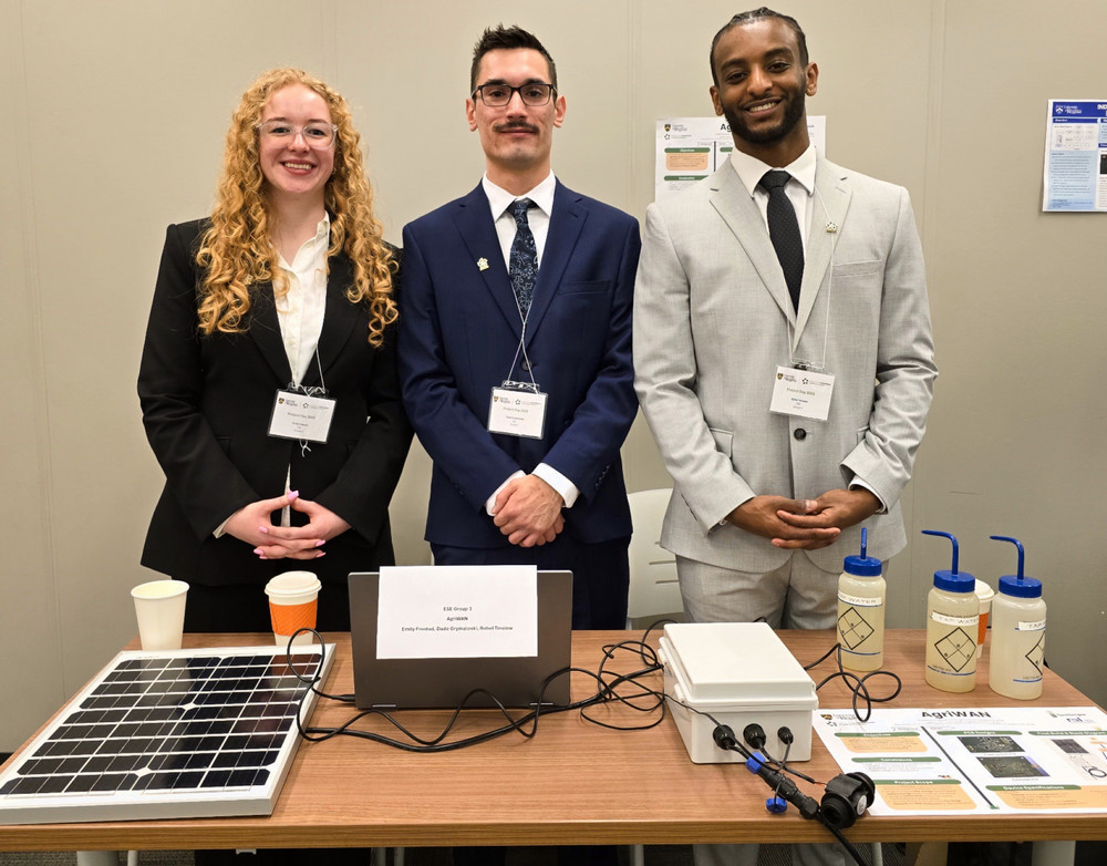 Three University of Regina Electronic Systems Engineering students smile confidently behind a display table 