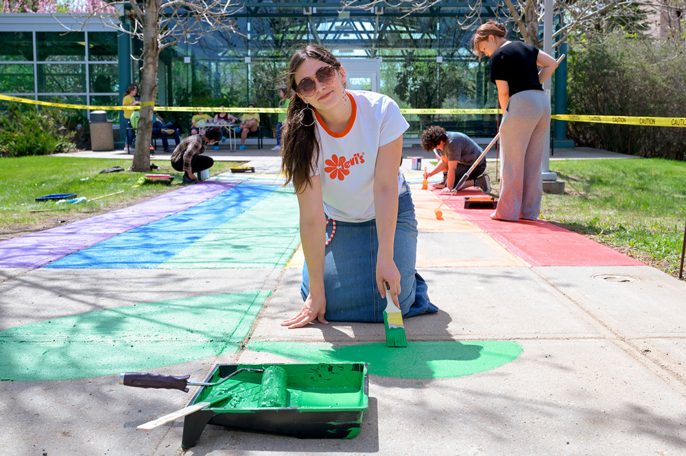 A student kneeling on a sidewalk paints a green section of a rainbow sidewalk while others paint additional colours in the background.