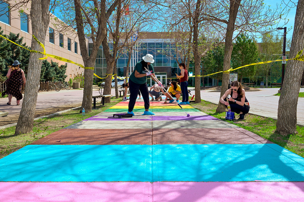 Several people paint rainbow stripes on a sidewalk outside the Riddell Centre.