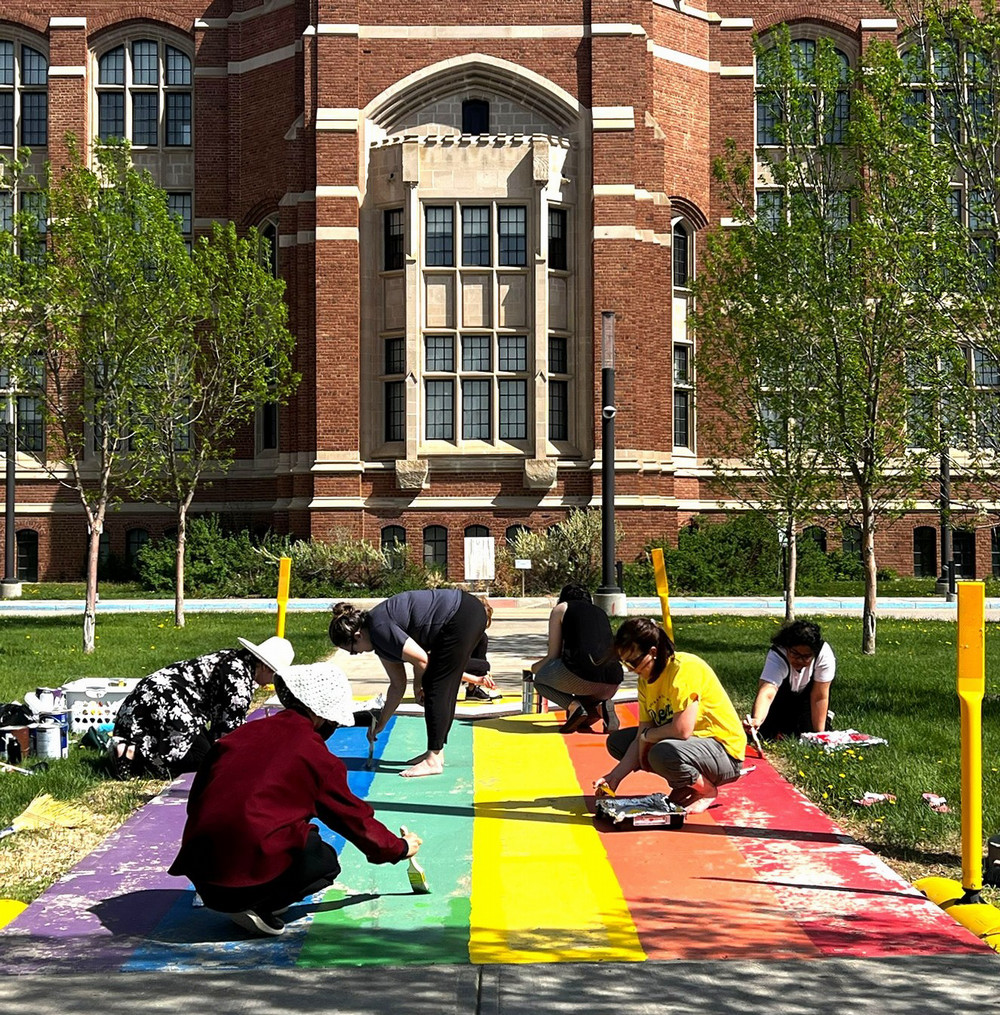 A group of people paint rainbow stripes on a sidewalk at the College Avenue Campus